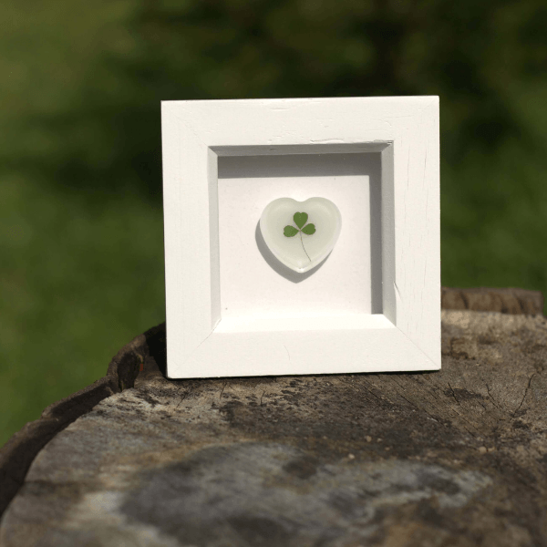 White framed heart-shaped resin with Hand foraged shamrock on a tree stump with a blurred green background